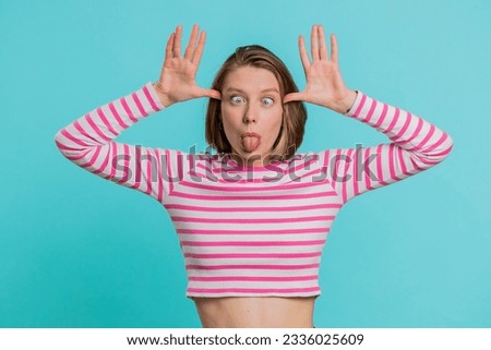 Funny comical playful young woman making silly facial expressions and grimacing, fooling around, showing tongue, idiotic expression. Pretty girl in crop top isolated alone on blue studio background