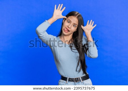 Funny comical playful young woman making silly facial expressions and grimacing, fooling around, showing tongue, idiotic expression. Girl isolated alone on blue studio background. People lifestyles