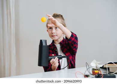 Funny Consentrating Little Boy Doing Home Science Project, Hovering Ping Pong Ball With A Fan. All Behind The Table.