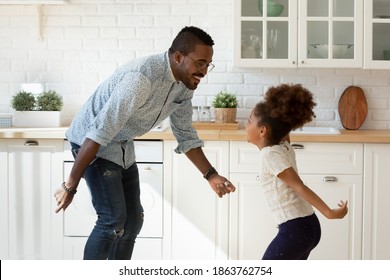 Funny Cooking With Dad. Happy Laughing Millennial Black Father Dancing Disco At Modern Designed Kitchen With Overjoyed Little Kid Daughter Jumping Listening To Music Enjoying Active Leisure Having Fun