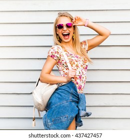 Funny cool cute beautiful hipster blonde girl posing against a white wall, bright casual wear, denim shirt, T-shirt, sunglasses, backpack, urban style, laughs, a broad smile, crazy emotions, joy