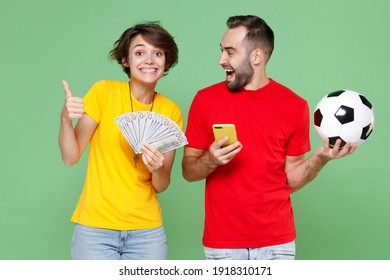 Funny couple friends sport family woman man football fan in t-shirt cheer up support favorite team with soccer ball using mobile phone hold money dollars showing thumb up isolated on green background