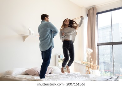 Funny couple jumping on bed in modern bedroom. Cheery happy woman and man in love celebrate relocation day, dance at cozy fashionable home. Tenancy, cohabitation, bank loan for young family concept