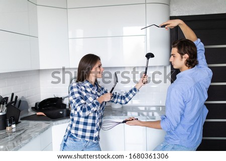 Funny couple pretending fight with utensils tools while cooking at home together, husband and wife having fun feeling playful holding kitchenware struggling in the kitchen preparing healthy food