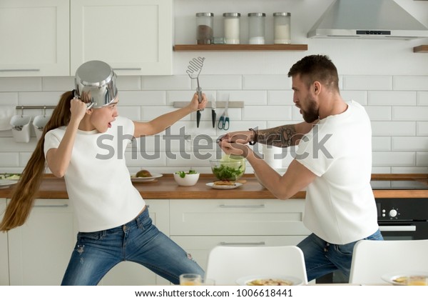 Funny couple pretending fight with utensils tools\
while cooking at home together, husband and wife having fun feeling\
playful holding kitchenware struggling in the kitchen preparing\
healthy food