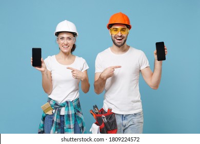 Funny couple woman man in hardhat glasses point index finger on mobile phone with blank empty screen isolated on blue background. Instrument accessories renovation apartment room. Repair home concept