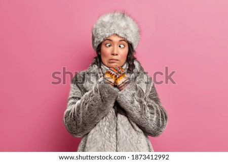 Funny crazy Eskimo woman makes grimace crosses eyes wears winter fur clothes has fun in studio poses against pink background. Arctic female plays fool dressed in coat and hat for frosty weather