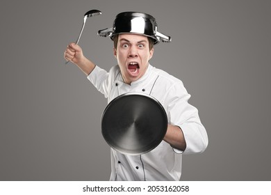 Funny crazy furious male chef in white uniform with saucepan on head and ladle and lid in hands pretending to fight, as knight on gray background