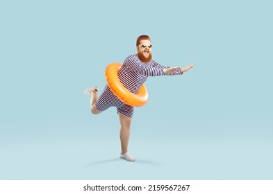 Funny crazy man with inflatable circle pretends to float on light azure background. Cheerful redhead bearded chubby guy in striped leotard having fun with swimming circle at waist. Full length. Banner
