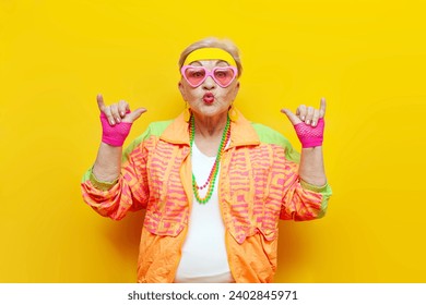 funny crazy old granny in sports hipster clothes shows youth gesture on yellow isolated background, elderly woman active lifestyle
