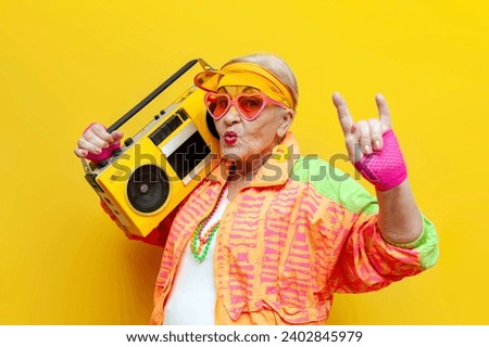 funny crazy old granny with tape recorder in sports hipster clothes listens to rock music on yellow background, elderly woman with record player at party and shows rock gesture