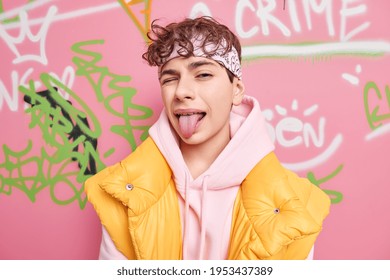 Funny curly haired teenage boy winks eye and stricks out tongue has fun poses against graffiti wall marks public places has playfull manners. Urban lifestyle and millennails generation concept