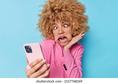 Funny curly haired woman goes crazy crosses eyes uses mouth expander has white even teeth makes grimace wears formal pink jacket takes selfie via modern smartphone isolated over blue background. - Shutterstock ID 2157768269