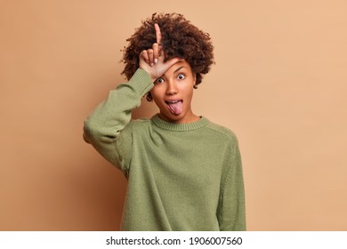 Funny curly haired young African American woman makes loser gesture sticks out tongue dressed in casual jumper isolated over brown background mocks at someone who lost bet. Body language concept