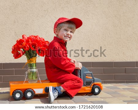 funny cute 4 years old boy in a red mechanic overalls and a cap sitting on a toy truck with large bouquet of tulips. Delivery concept, little courier,  gift for women, with love for mommy. Copy space