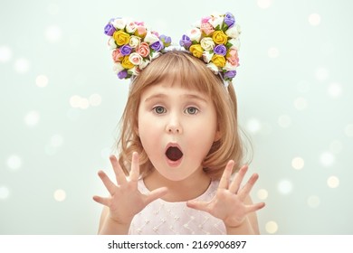 A funny and cute 4-year old girl in pink summer dress and floral hair rim stands with big surprise on her face holding hands in front of her. Light background with shining lights. Children's fashion. 