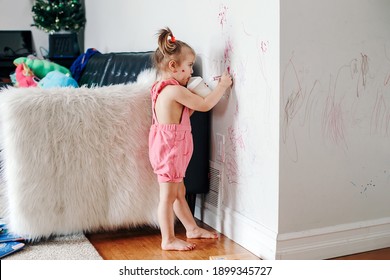 Funny cute baby girl drawing and marker wall at home  Toddler girl child and milk bottle playing at home  Authentic candid childhood lifestyle moment  Young artist painting wall in living room