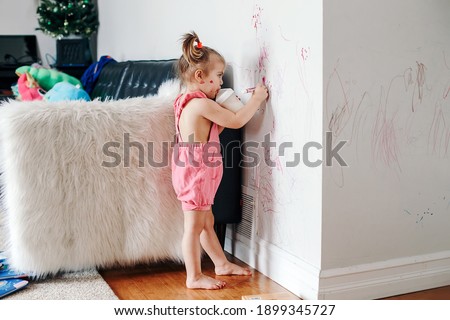 Funny cute baby girl drawing with marker on wall at home. Toddler girl child with milk bottle playing at home. Authentic candid childhood lifestyle moment. Young artist painting on wall in living room