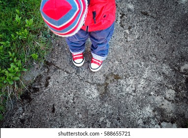 Funny Cute Boy Looking Onto Own Feet. Baby Standing On Saphalt In Red Shoes