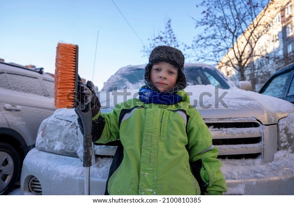 Funny cute Caucasian boy cleans the car from snow\
with a brush and a\
scraper