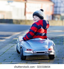 Funny Cute Child In Red Jacket Driving Big Vintage Old Toy Car And Having Fun, Outdoors. Kids Leisure On Cold Day In Winter, Autumn Or Spring.