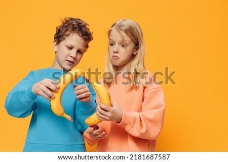funny, cute children, a boy and a girl stand on a blue background in bright clothes and holding bananas in their hands are fooling around, connecting fruits in the shape of a circle