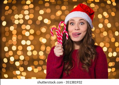 Funny cute girl in santa claus hat with christmas lollypops over shining background