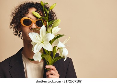 Funny cute handsome tanned curly man in trendy sunglasses hold lilies near face posing isolated on over beige pastel background. Fashion New Collection offer. Retro style concept. Free place for ad