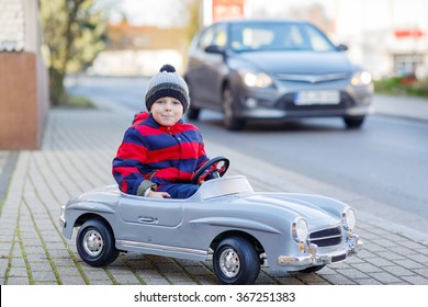 Funny Cute Kid Boy In Red Bright Jacket Driving Big Vintage Old Toy Car And Having Fun, Outdoors. Active Leisure For Kids On Cold Day In Winter, Autumn Or Spring.