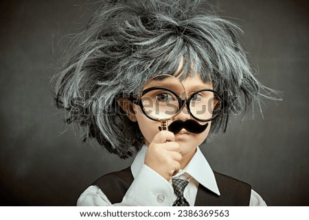 Funny cute kid in glasses, with a mustache and in a wig, like little professor looks through a magnifier. Against the background of a black chalkboard. Little Scientists. Education.