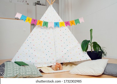 Funny cute little kid's feet sticking out from under a hut, made with sticks and bedsheets. Stay at home flag garland hanging across the room overhead.