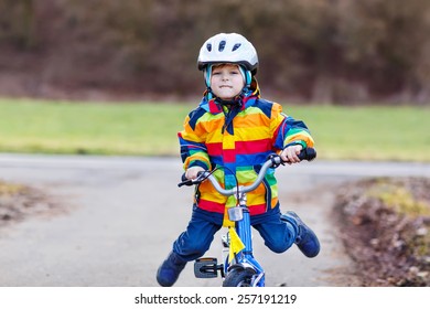 Funny Cute  Preschool Kid Boy In Safety Helmet And Colorful Raincoat Riding His First Bike And Having Fun On Cold  Day, Outdoors. Active Leisure With Children In Winter, Spring Or Autumn.