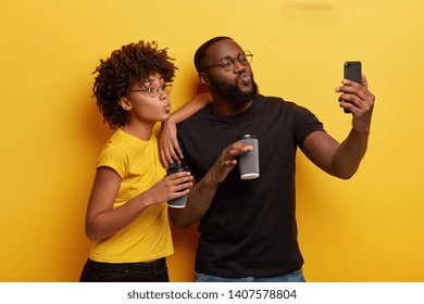 Funny Dark Skinned Couple Pouts Lips At Camera Of Cell Phone, Make Selfie Portrait, Drink Coffee To Go From Disposable Cups, Wear Black And Yellow T Shirts, Round Spectacles, Pose Indoor, Have Fun