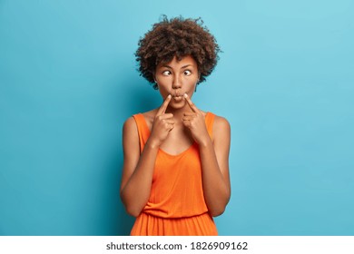 Funny Dark Skinned Woman Crosses Eyes And Makes Fish Lips Funny Grimace Dressed In Fashionable Dress Isolated On Blue Background. Ridiculous Hilarious Idiotic Afro American Female Model Indoor