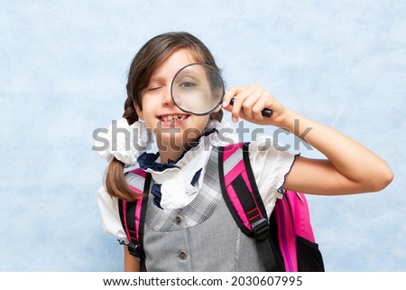 Funny dark-haired schoolgirl with pigtails. Fooling around on a blue background. With a magnifying glass. Portrait. Place for your text.