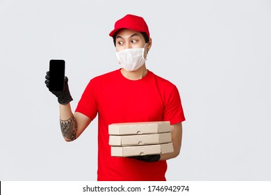 Funny Delivery Guy In Red Cap, T-shirt, Holding Pizza Boxes And Show Smartphone Screen. Asian Courier Confirm Order Info With Client, Bring Food To People Home During Quarantine, Wear Medical Mask