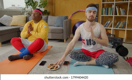 Funny diverse male friends meditating together at home training. Cunning bearded freak guy eating sweet cookie without being seen. Fun and entertainment.