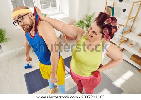 Funny dominant female fat woman leader raising her boyfriend in sportswear by hand dominating him during doing sport exercises with dumbbells at home. Domination, submission and violence concept.