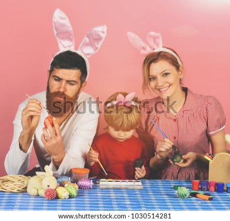 funny easter egg Mother, father and daughter preparing for Easter. Man with beard, woman and kid with happy faces painting eggs on pink background. Family members wearing cute bunny ears.