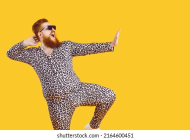 Funny eccentric overweight man dressed in leopard print pajamas dancing and fooling around on orange background. Cheerful bearded fat man in sunglasses having fun at copy space. Web banner.