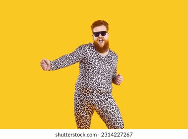Funny eccentric overweight man in pajamas with leopard print having fun, dancing and fooling around. Millennial red-bearded fat man in pajamas and black glasses dances isolated on orange background.