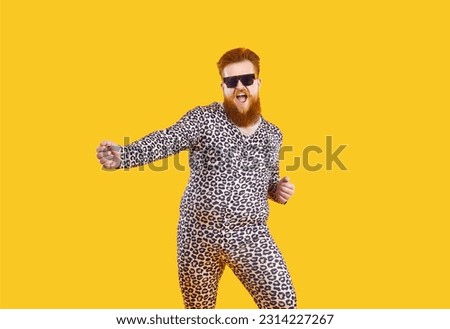 Funny eccentric overweight man in pajamas with leopard print having fun, dancing and fooling around. Millennial red-bearded fat man in pajamas and black glasses dances isolated on orange background.
