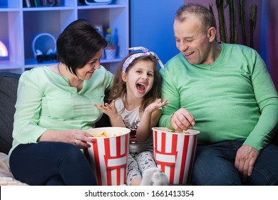 Funny Elder Old Family And Little Kid Granddaughter Sits On Sofa And Watching Tv Eating Popcorn.