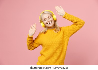 Funny elderly gray-haired blonde woman lady 40s 50s years old in yellow casual sweater listening music with headphones keeping eyes closed dancing isolated on pastel pink background studio portrait