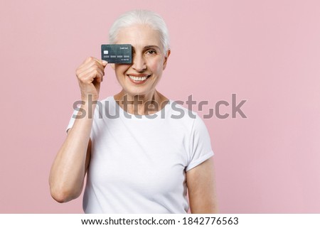 Funny elderly gray-haired female woman 60s 70s in white casual blank design t-shirt posing covering eye with credit bank card looking camera isolated on pastel pink color background studio portrait