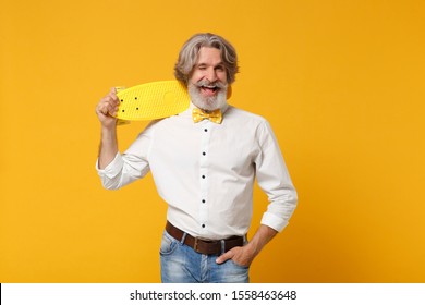 Funny Elderly Gray-haired Mustache Bearded Man In White Shirt And Bow Tie Isolated On Yellow Orange Background Studio Portrait. People Lifestyle Concept. Mock Up Copy Space. Hold Skateboard, Blinking