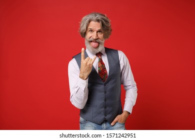 Funny elderly gray-haired mustache bearded man in classic shirt vest tie isolated on red wall background. People lifestyle concept. Mock up copy space. Showing tongue, depicting heavy metal rock sign