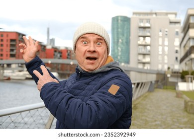 funny elderly man in jacket points to tower, modern buildings in Frankfurt, 30-story skyscraper Westhafen Tower in Gutleitviertel district, concept of winter city walks, international tourism