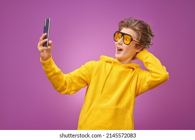 A funny emotional boy teenager in bright yellow hoodie talking gaily via video call using his smartphone. Bright purple background. Adolescents, lifestyle and communications. Modern teenagers. 