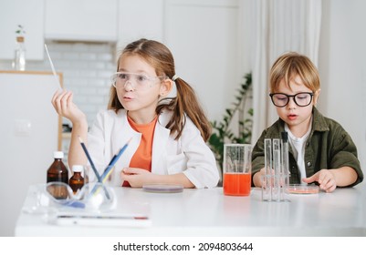 Funny Emotional Little Girl In White Robe Holding A Pipette, Next To Her Boy Picks A Dish With Red Chemical Liquid. Doing A Home Science Project Behind The Table.